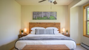 A bedroom with a wooden headboard, a large bed with white bedding, two bedside tables with lamps, a bench, and a painting of a forest on the wall.