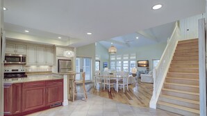 A spacious kitchen with modern appliances, a dining area with white chairs, and a wooden staircase leading to a living area. - 233 Beachside Home- Large 4 Bedroom Homes with Heated Community Pool (Hilton Head Island)