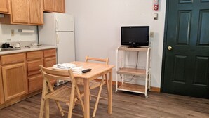 A compact kitchen with wooden cabinets, a white refrigerator, a dining table with chairs, a television on a stand, and a door with a fire alarm. - Hotel Style Lodge Room w/Kitchenette (Almont)