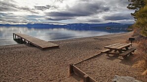 Una playa arenosa con un muelle de madera y una mesa de picnic, con vistas a un lago tranquilo y montañas al fondo.