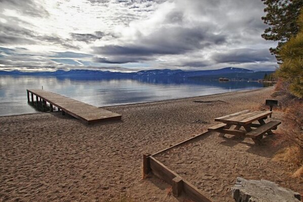 A sandy beach with a wooden dock and picnic table, overlooking a calm lake with mountains in the background.