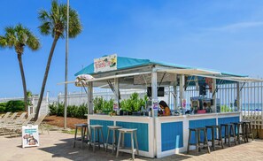 A beachside food kiosk with a blue awning, a menu board, and a sign for a weekly shrimp boil.