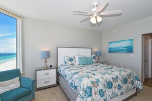 Ein Schlafzimmer mit einem Bett, Nachttischen, einem Deckenventilator, einem Fenster mit Blick auf den Strand und einem Bild an der Wand.