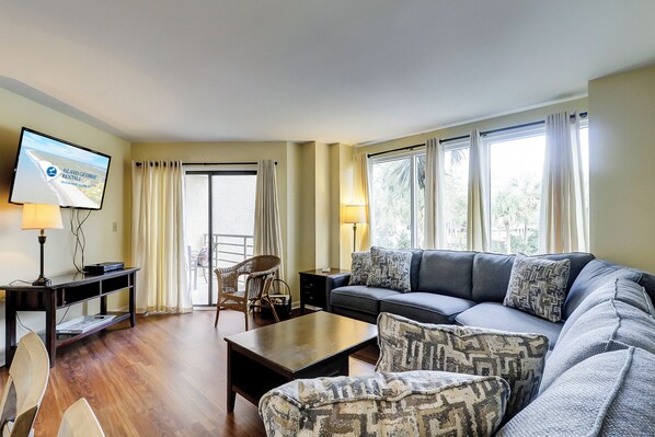 A living room with a sofa, armchairs, a coffee table, a TV on a stand, and a view of the outdoors through large windows.