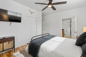 Une chambre à coucher comprenant un lit, un ventilateur de plafond, un téléviseur fixé au mur et une porte donnant accès à une salle de bain.