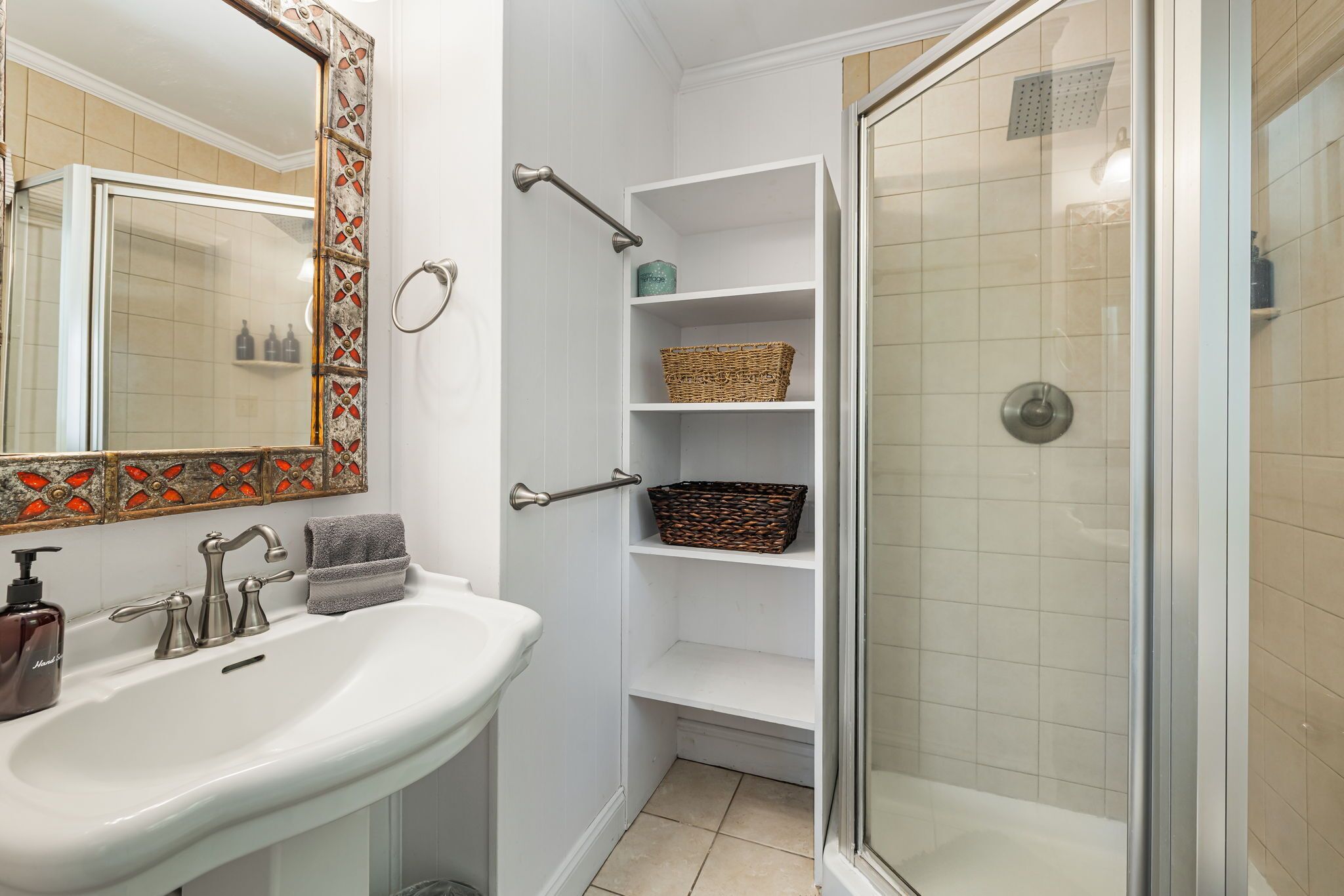 Un baño con un lavamanos blanco, un espejo con marco decorativo, una ducha con puerta de cristal y un estante con cestas.