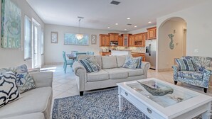 Una sala de estar amplia con sofá, sillón y mesa de centro, una cocina con armarios de madera y un comedor con lámpara de araña.