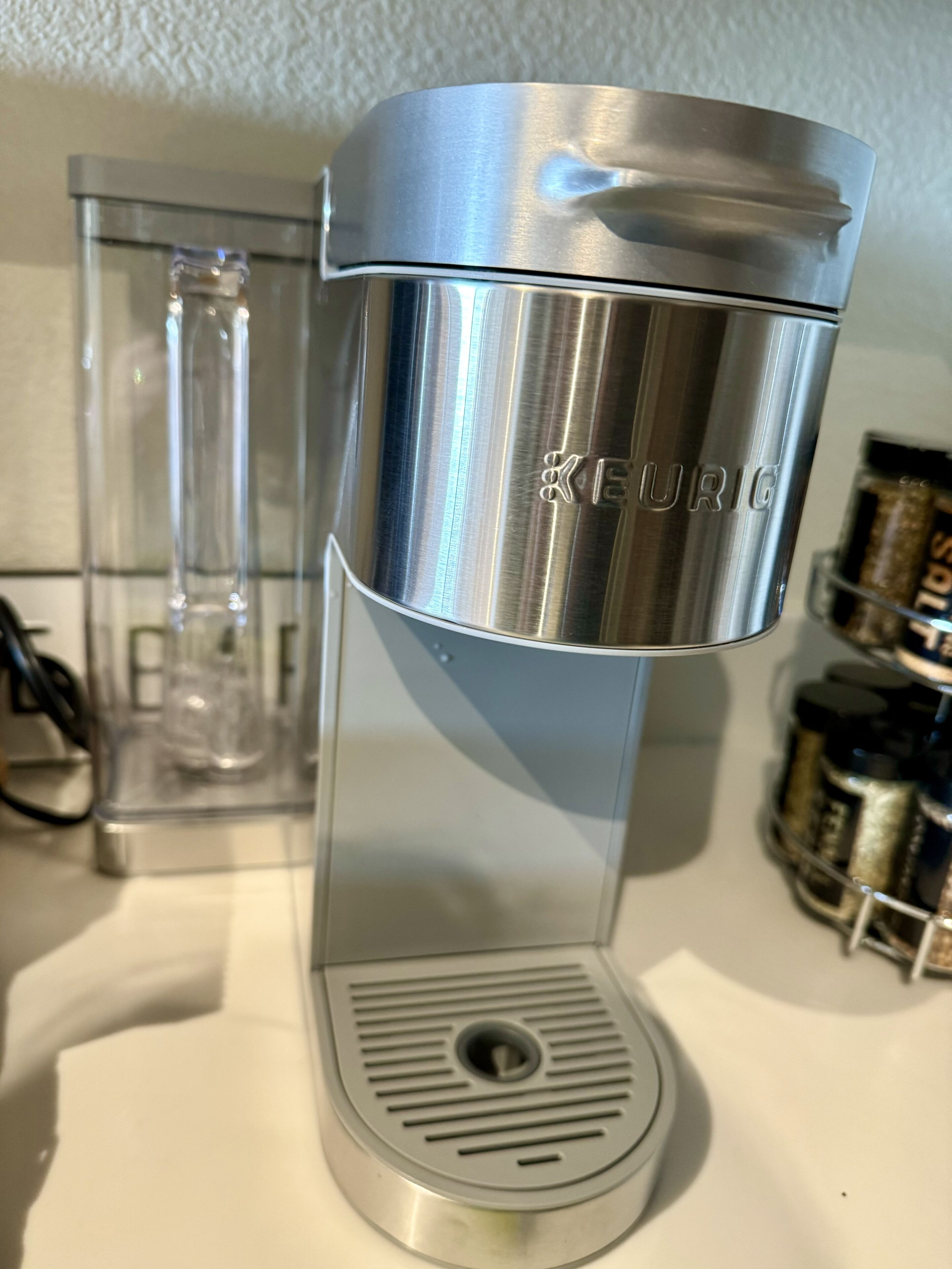 A Keurig coffee maker on a white countertop with a clear glass carafe and a spice rack in the background.