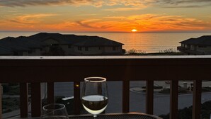 En balkon med to glas hvidvin, udsigt til en solnedgang over havet med huse i baggrunden.
