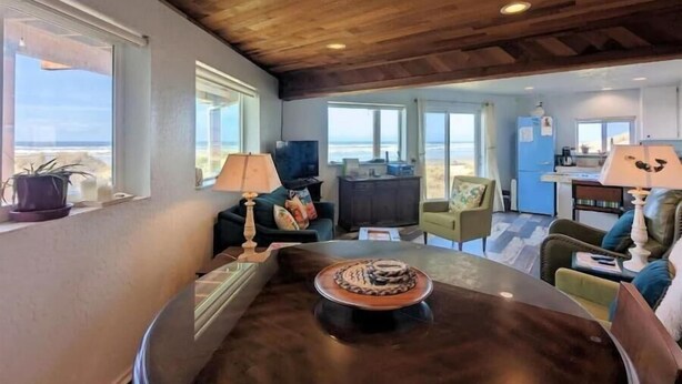 Amplia sala de estar con techo de madera, grandes ventanales con vista al océano, mesa de comedor con un centro decorativo y zona de cocina visible a través de un umbral abierto.