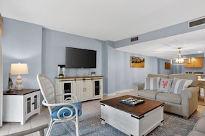 A living room with a flat-screen TV, a white cabinet, a sofa, a coffee table, and a dining area with a chandelier.