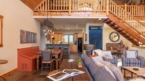 A spacious living area with a high ceiling, a dining area, and a kitchen. There is a staircase leading to an upper level.