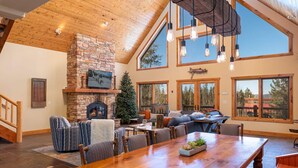 A spacious living area with a large wooden dining table, comfortable seating, a stone fireplace, and large windows offering views of the outdoors.