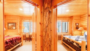 Ein Blockhaus-Interieur mit einem zentralen Holzbalken, einem Bett mit Karomuster-Bettwäsche, einem Schreibtisch mit Stuhl und einem Fenster mit Blick auf Bäume.