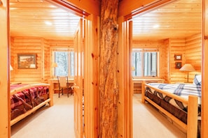 Ein Blockhaus-Interieur mit einem zentralen Holzbalken, einem Bett mit Karomuster-Bettwäsche, einem Schreibtisch mit Stuhl und einem Fenster mit Blick auf Bäume.