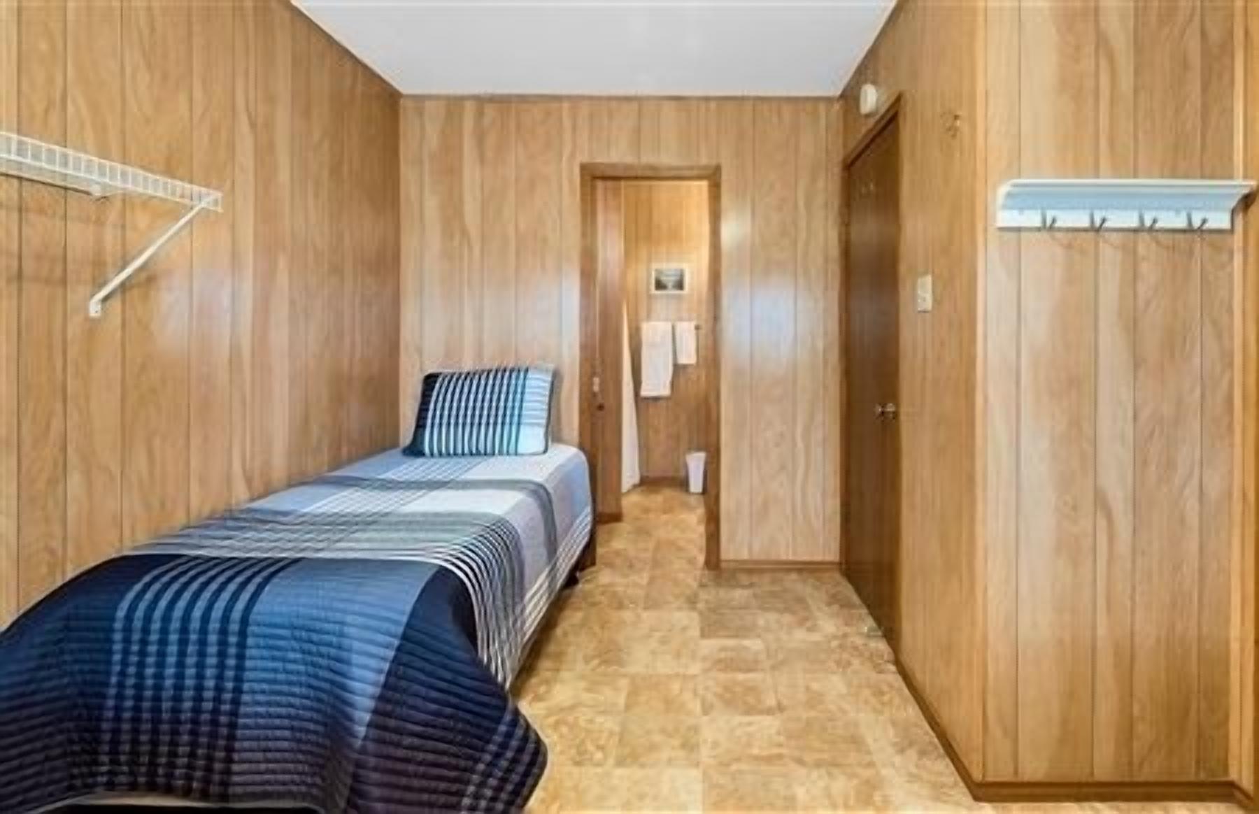 A small, compact room with wooden walls and a tiled floor. It includes a bed with a striped bedspread, a wardrobe, and a small hallway leading to a door.