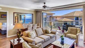 Ein geräumiges Wohnzimmer mit einem großen Sofa, zwei Sesseln und einem Glastisch. Es gibt einen Esstisch mit Stühlen, ein großes Fenster mit Meerblick und einen Balkon mit Gartenmöbeln.