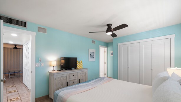 Une chambre à coucher avec un lit, un ventilateur de plafond, un téléviseur posé sur une commode et une porte donnant sur une autre pièce.