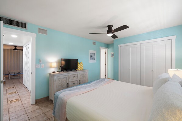 A bedroom with a bed, a ceiling fan, a television on a dresser, and a door leading to another room.