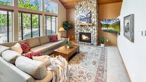 A cozy living room with a stone fireplace, a sofa, a coffee table, and a large window with blinds. - Unique Covered Silo Lounge | Fire-Pit Gatherings+ EV Ready & 1PM Check-IN/OUT (ronald)