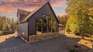 A modern cabin with a stone foundation, large windows, and a sloped roof, surrounded by trees and a gravel driveway. - Unique Covered Silo Lounge | Fire-Pit Gatherings+ EV Ready & 1PM Check-IN/OUT (ronald)