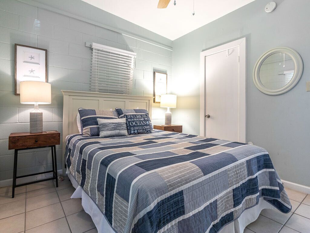 Une chambre à coucher comprenant un lit, deux tables de chevet, une commode, une fenêtre avec des stores, une porte et un miroir fixé au mur.