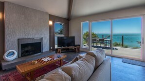 A modern living room with a fireplace, a television, and a large sliding glass door leading to a balcony with a view of the sea. - Treasure Cove-Hot Tub, Ocean Views & Near Beach (Trinidad)