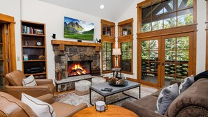 A cozy living room with a stone fireplace, leather sofas, a coffee table, and large windows with wooden frames. - Roomy, Luxurious Ski-In/Ski-Out w/ Views, Hot Tub! Bachelor Gulch, Beaver Creek (Edwards)