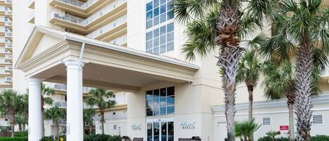 A hotel entrance with a covered walkway, palm trees, and a multi-story building in the background.