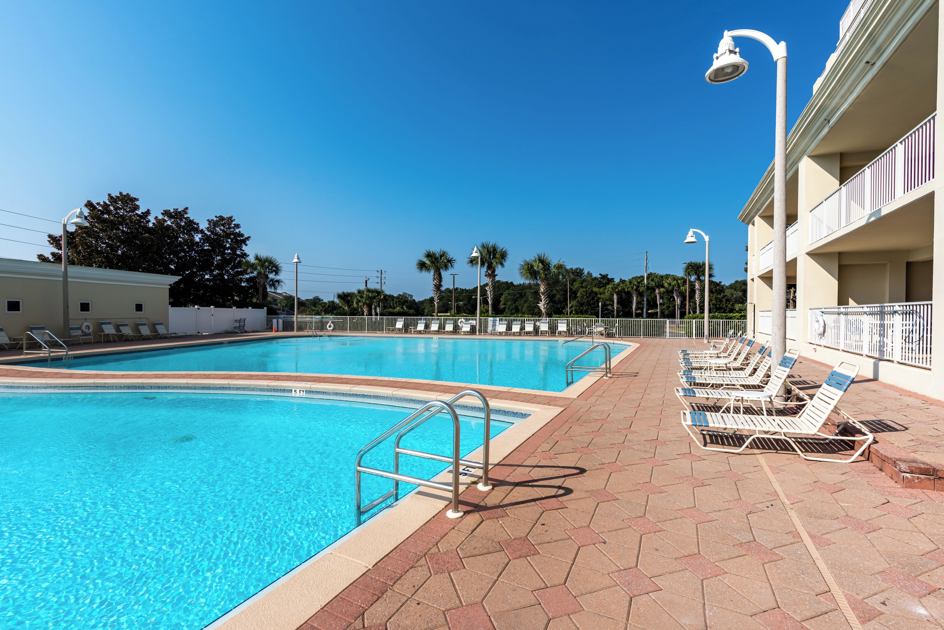 Ein Schwimmbecken mit gefliestem Sonnendeck, Liegestühlen und einem Gebäude mit Balkonen im Hintergrund.