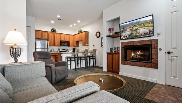 A living room with a fireplace, a television mounted above it, and a kitchen in the background.