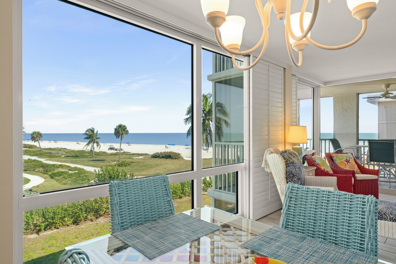 A room with a large window overlooking a beach, wicker furniture, and a ceiling fan.