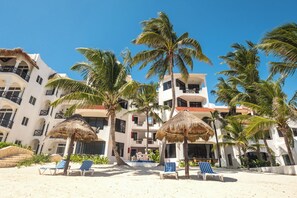 A beachfront hotel with multiple balconies, palm trees, and thatched umbrellas.