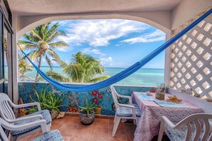 A patio with a hammock, chairs, and a table with a view of the ocean and palm trees.
