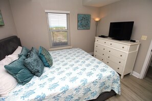 Une chambre à coucher comprenant un lit, une commode, un téléviseur et une fenêtre avec des stores.