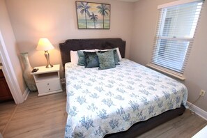 Une chambre à coucher avec un lit, une table de chevet, une lampe, un tableau et une fenêtre avec des stores.