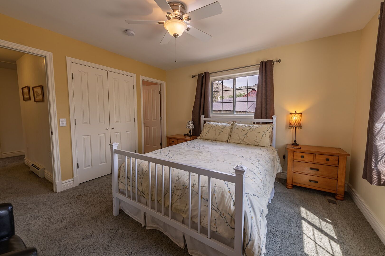 A bedroom with a bed, a nightstand, a lamp, a window with curtains, a ceiling fan, and a door.