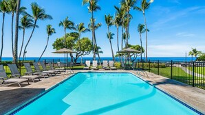 A swimming pool with lounge chairs and umbrellas, surrounded by palm trees and a view of the ocean.