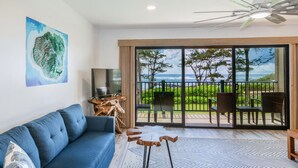 A modern living room with a sofa, a TV, and a large window overlooking a balcony with outdoor furniture. - Pono Kai A-201  Air conditioned,  Oceanfront Condo on the Coconut Coast (Kapaa)