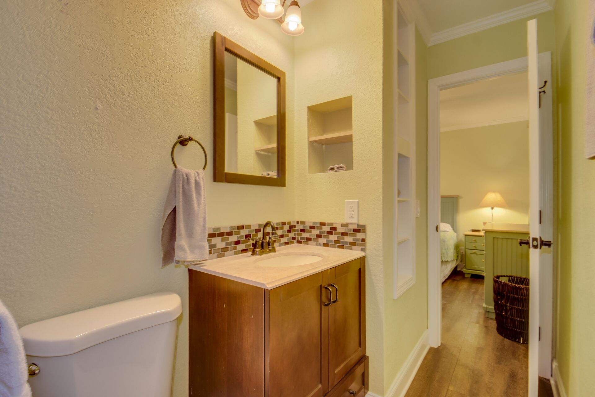 A bathroom with a toilet, sink, and mirror. There is a towel hanging on a ring, and a small shelf above the sink. The walls are painted in a light color, and there is a wooden cabinet below the sink.