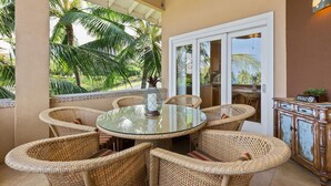 A covered outdoor area with a round glass table and wicker chairs, a white door, and a view of palm trees.