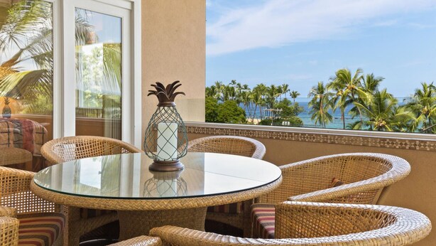 A round glass table with wicker chairs, a decorative vase, and a view of palm trees and the ocean.