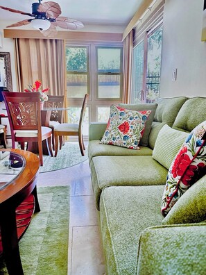 A living room with a green sofa, a dining table with chairs, a ceiling fan, and large windows.