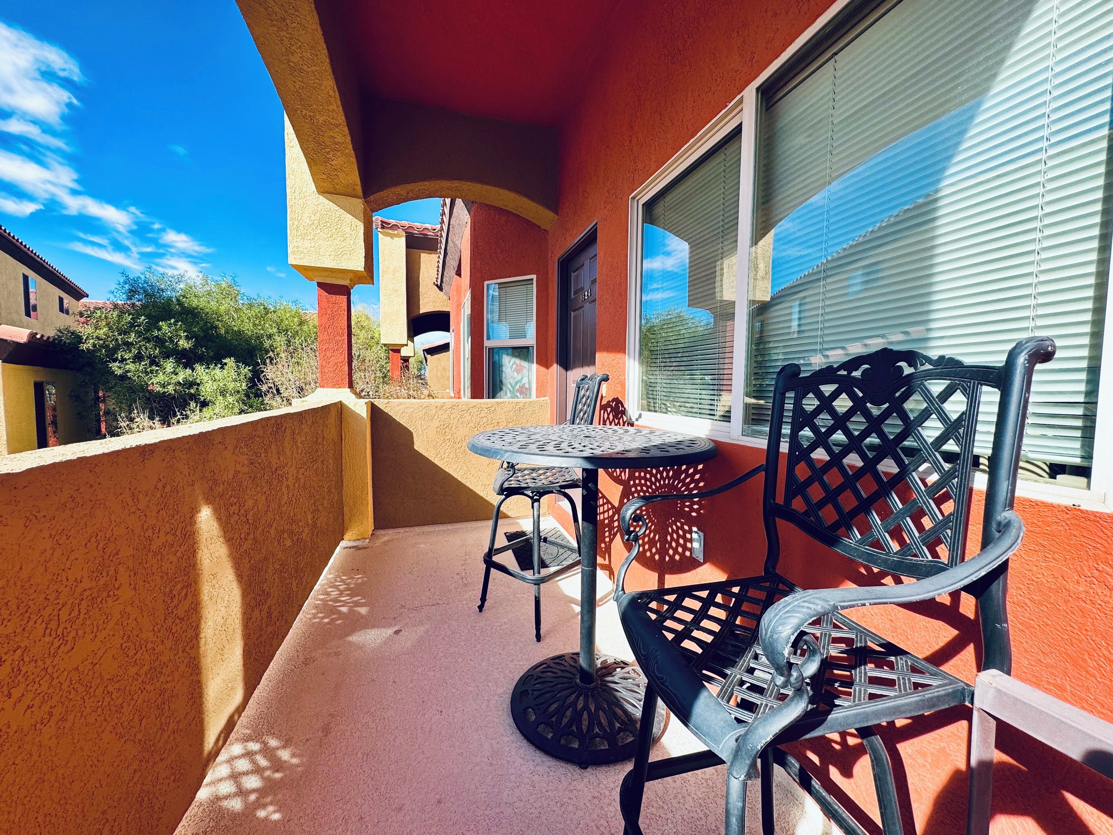 A balcony with a round table and two chairs, a red wall, and a window with blinds.