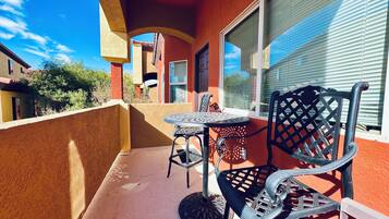 A balcony with a round table and two chairs, a red wall, and a window with blinds.