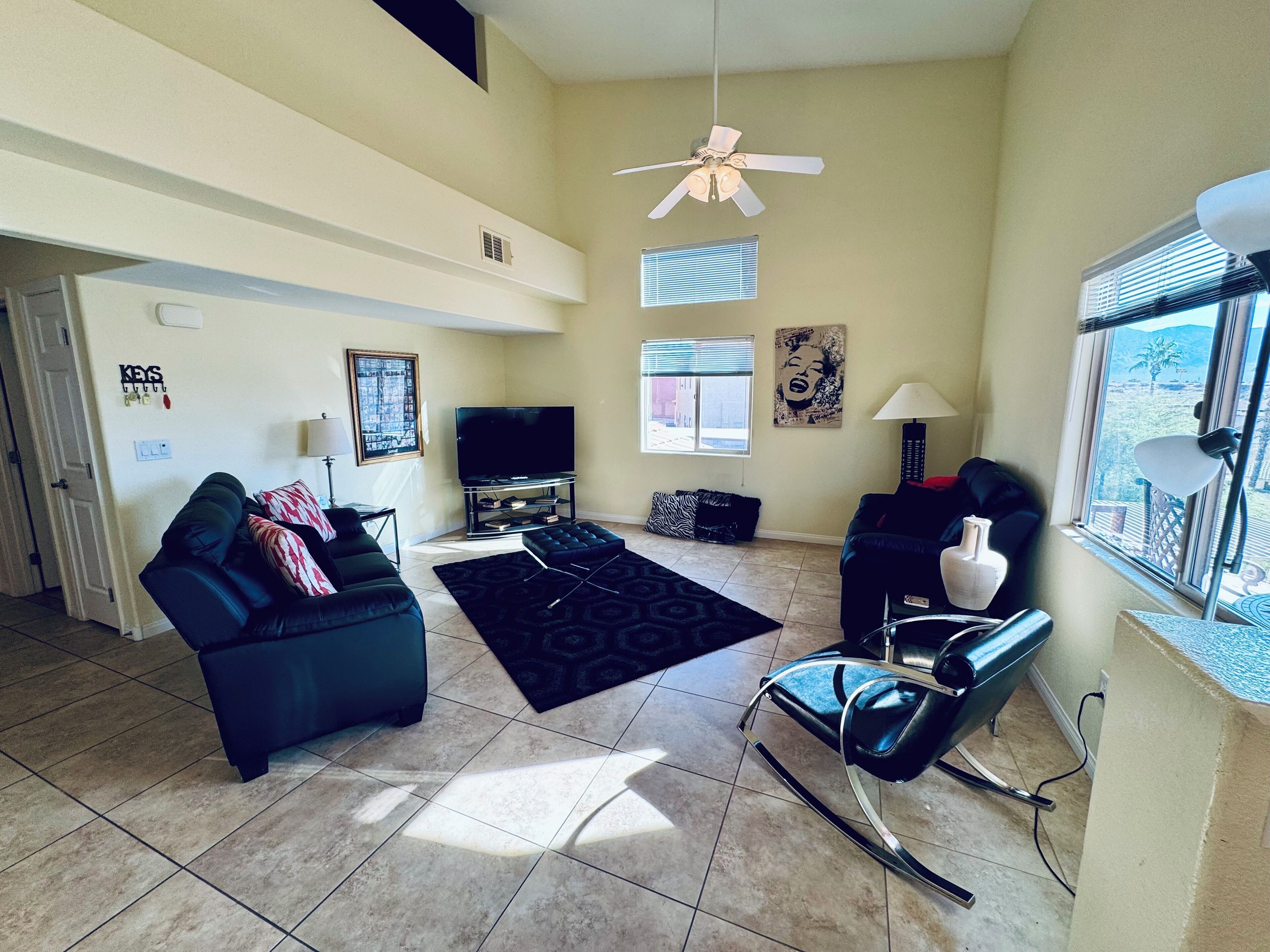 A living room with a sofa, ottoman, and a chair. There is a television, a window with blinds, and a ceiling fan.