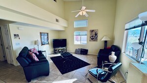A living room with a sofa, ottoman, and a chair. There is a television, a window with blinds, and a ceiling fan. - Sunset Sands by Mesquite Vacation Rentals: Pet Friendly near Wolf Creek with $510 in free daily activities (Mesquite)