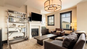 A living room with a fireplace, a television mounted above it, a sofa, a coffee table, and a bookshelf. - Modern Mountainside Townhome, Natural Light, Outdoor Recreation + Hot Tub! (Park City)