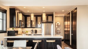 A modern kitchen with dark cabinetry, a central island with stools, a refrigerator, and pendant lighting. - Modern Mountainside Townhome, Natural Light, Outdoor Recreation + Hot Tub! (Park City)
