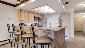 A kitchen with wooden cabinets, a microwave, a refrigerator, and a bar area with stools. - Perfect Downtown Retreat with ALL NEW APPLIANCES AS OF FALL 2025! (Breckenridge)